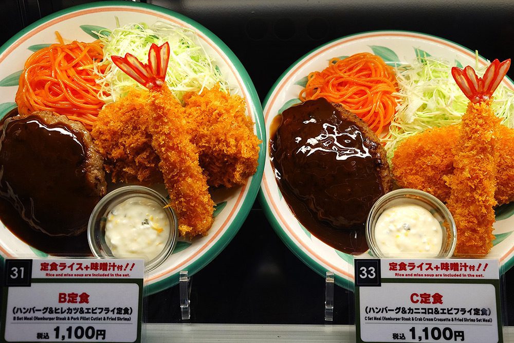 Close-up of two numbered food replica price tags — number 31 B Set Meal and number 33 C Set Meal, both at 1,100 yen, with red accent panels signalling that rice and miso soup are included