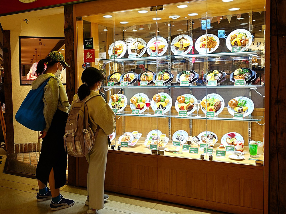 Cafe window display with food replicas and price tags, two pedestrians examining the menu before entering