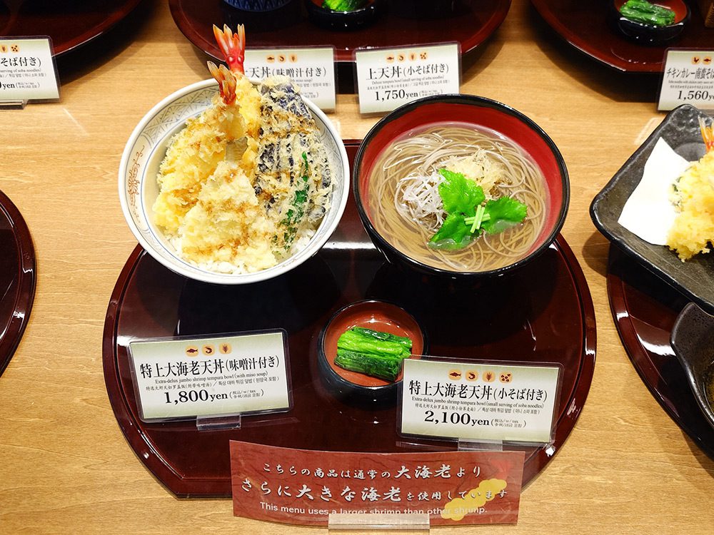 Two price tags side by side for the same tempura dish — extra-jumbo shrimp tempura bowl with miso soup at 1,800 yen and with small soba serving at 2,100 yen