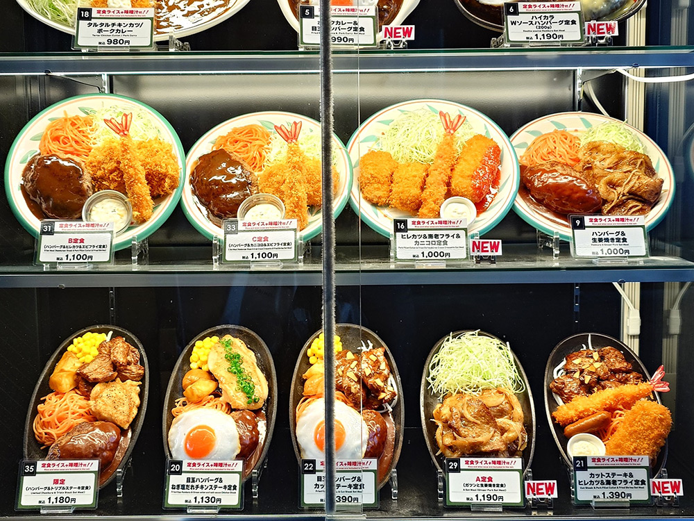 Food replica display with numbered price tags and NEW flags — every dish carries a digit allowing guests to order by number, while red NEW markers highlight new menu items