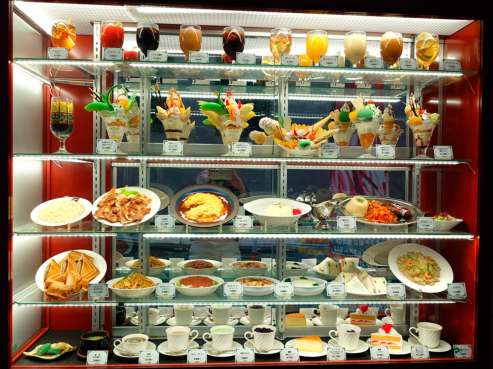Drink replicas on a side shelf beside desserts in a restaurant drink display