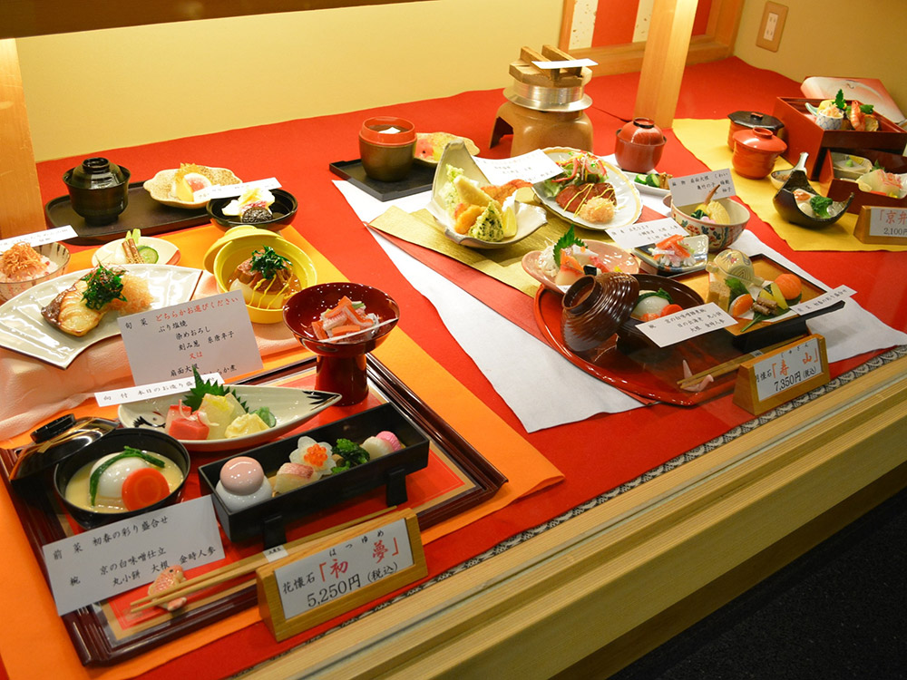 Japanese restaurant window display with red backdrop, kaiseki set replicas and price cards