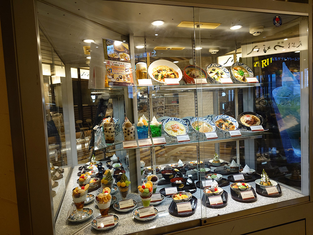 Glass display case with parfaits and noodle bowl replicas reflected in a mirrored side wall